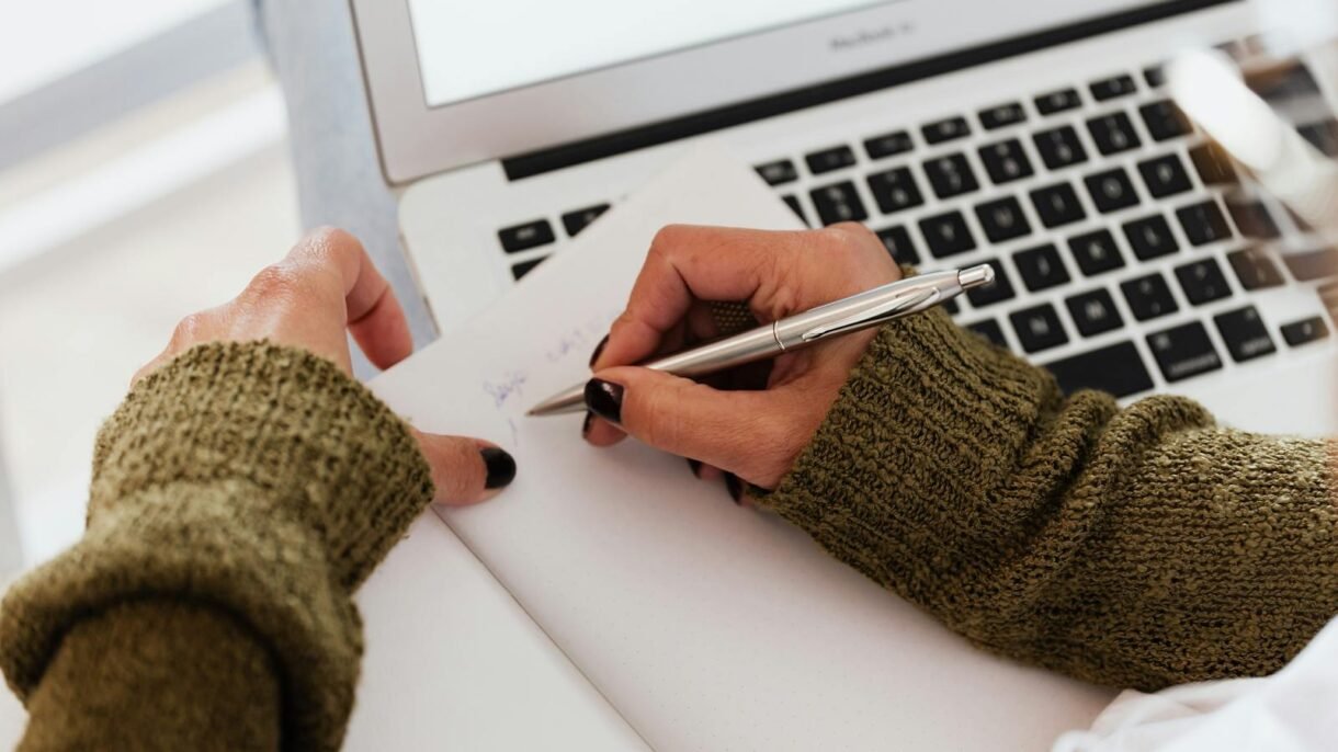 From above crop unrecognizable female in casual warm sweater taking notes in opened notebook with stylish silver pen while using modern netbook